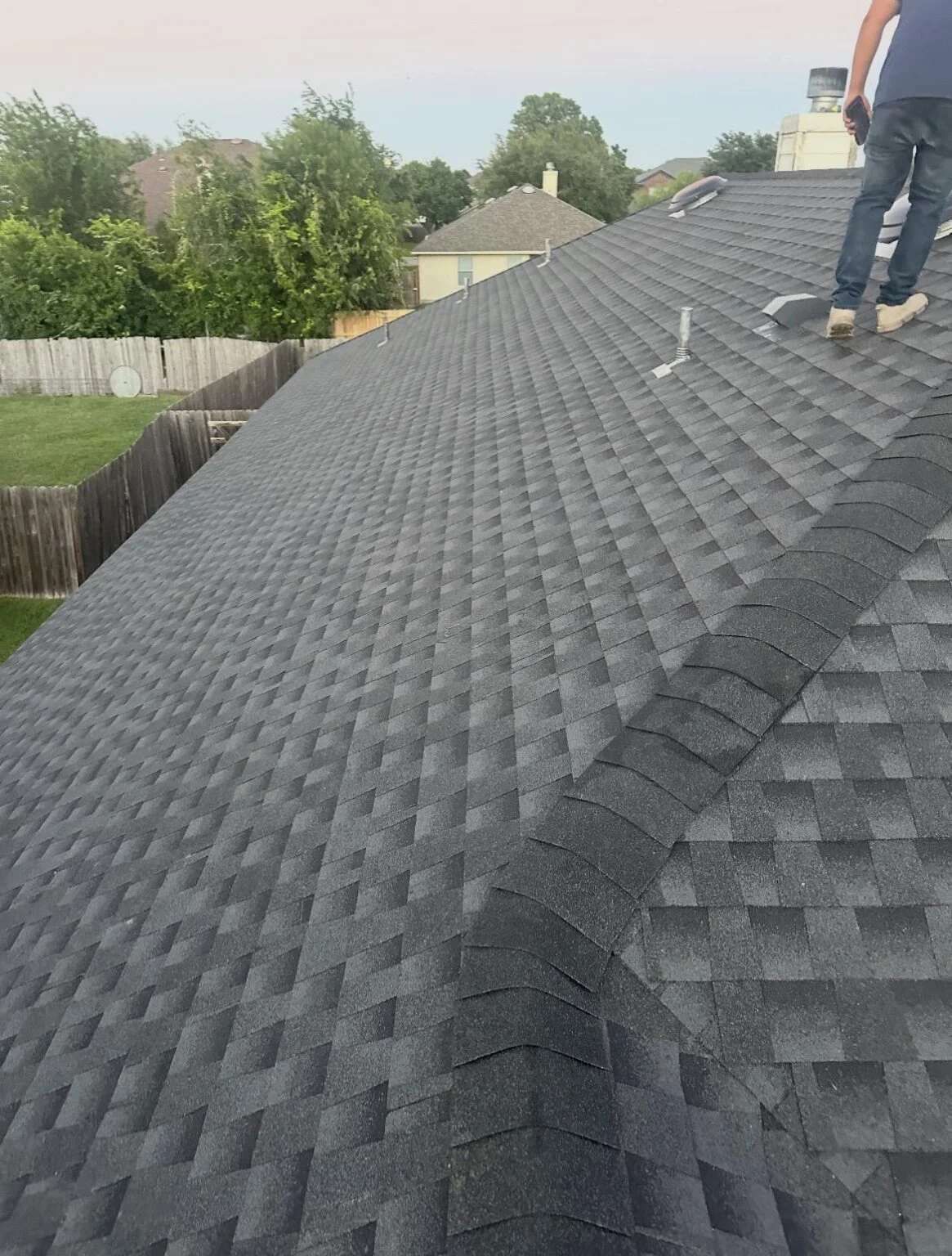 Person standing on a sloped shingled roof with tools and vent pipes nearby, overlooking a suburban backyard.