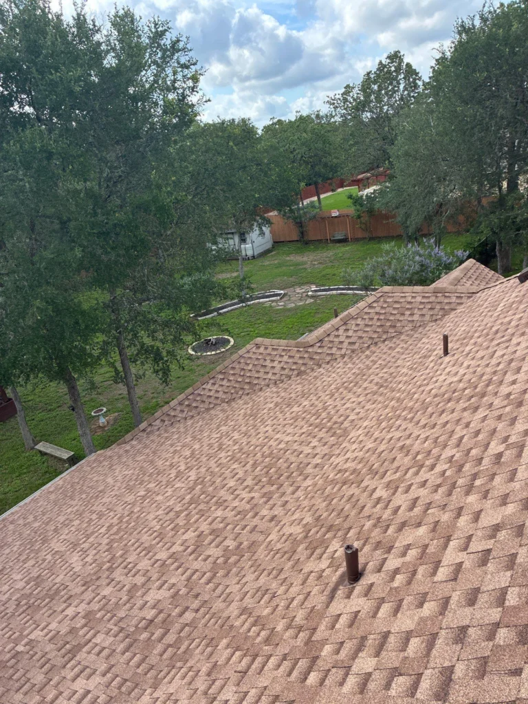 driftwoodbuildersroofingcom-asphalt-shingle-full-roof-replacement-in-bastrop-tx-asphalt-shingle-full-roof-replacement-in-bastrop-tx-1-768x1024 - Driftwood Builders Roofing - Driftwood Builders Roofing View of a brown shingled roof with a vent pipe, overlooking a backyard with trees, a circular fire pit, a stone path, and a wooden fence beyond the lawn.