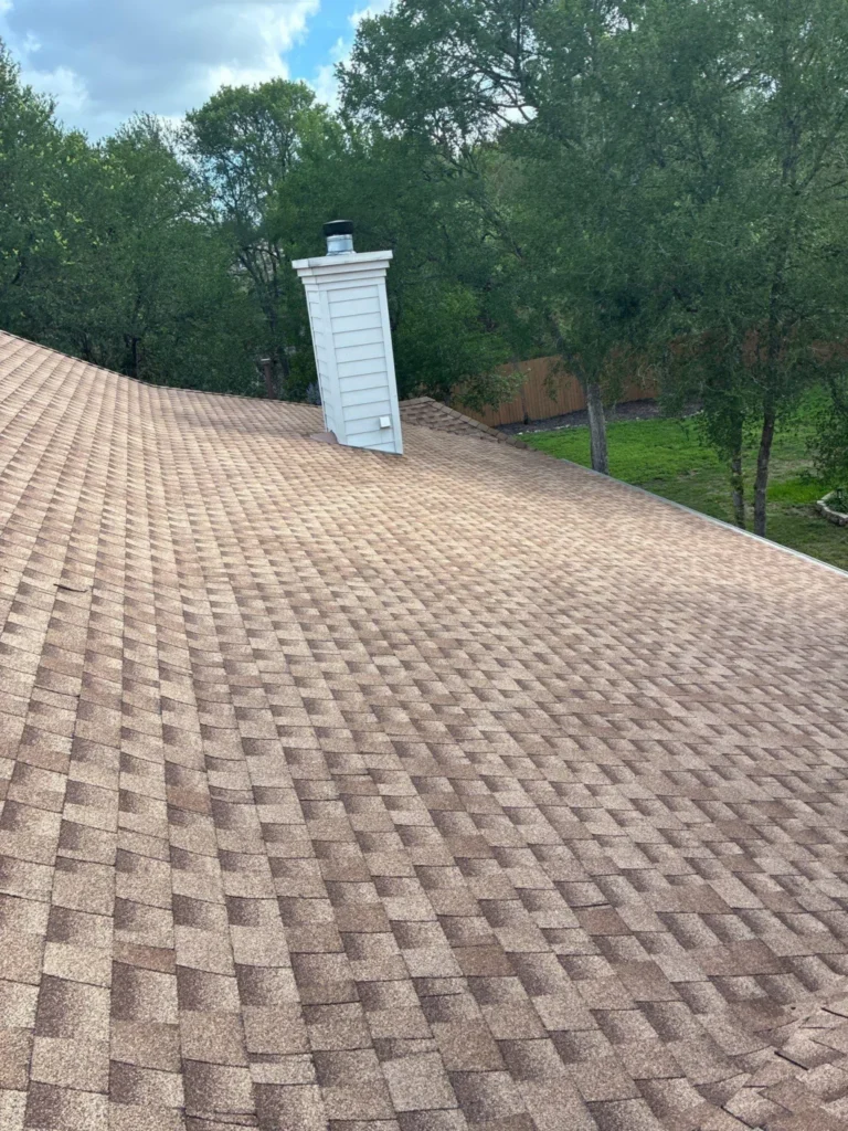 Brown shingled roof with a white chimney vent near the center and green trees in the background.