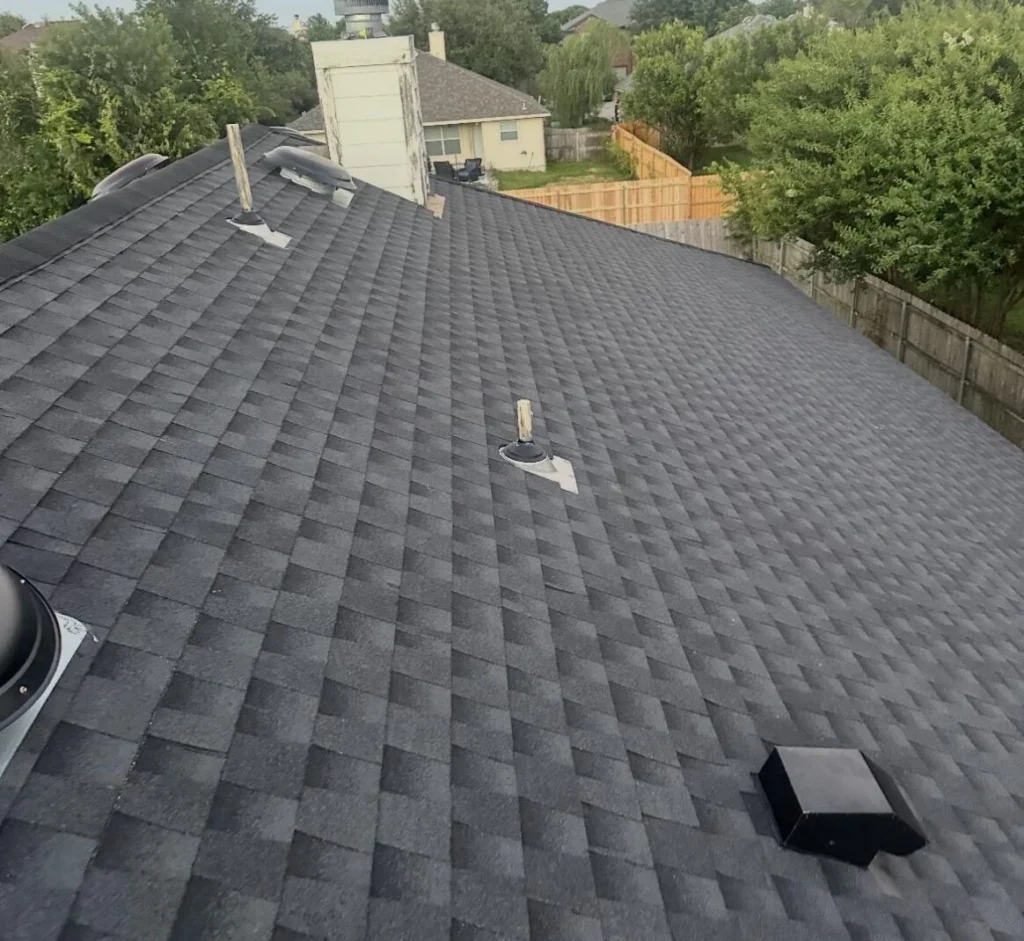 Aerial view of a dark gray shingle roof with vents and a chimney, with neighboring yards and trees in the background.