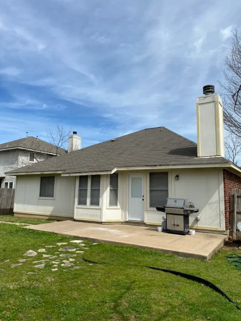 Backyard of a single-story cream house with a white door, large windows, and a stainless steel grill on a concrete patio under a blue sky.