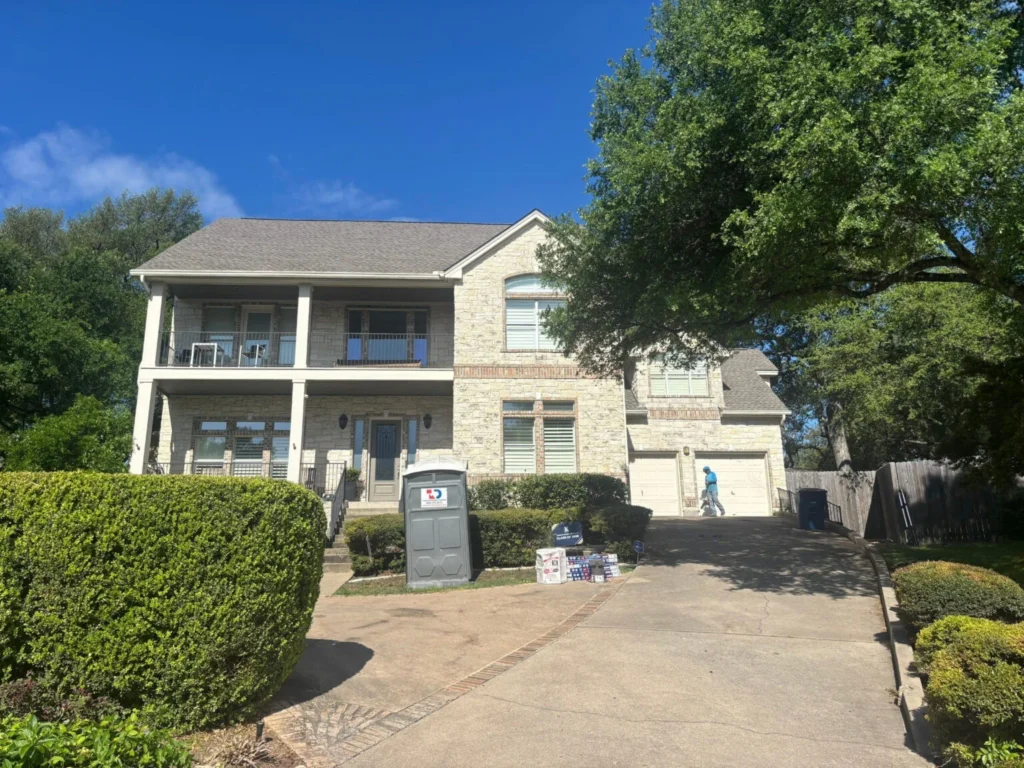 Two-story stone house with a front porch and balcony, manicured hedges, and a person walking up the sunlit driveway on a clear day.