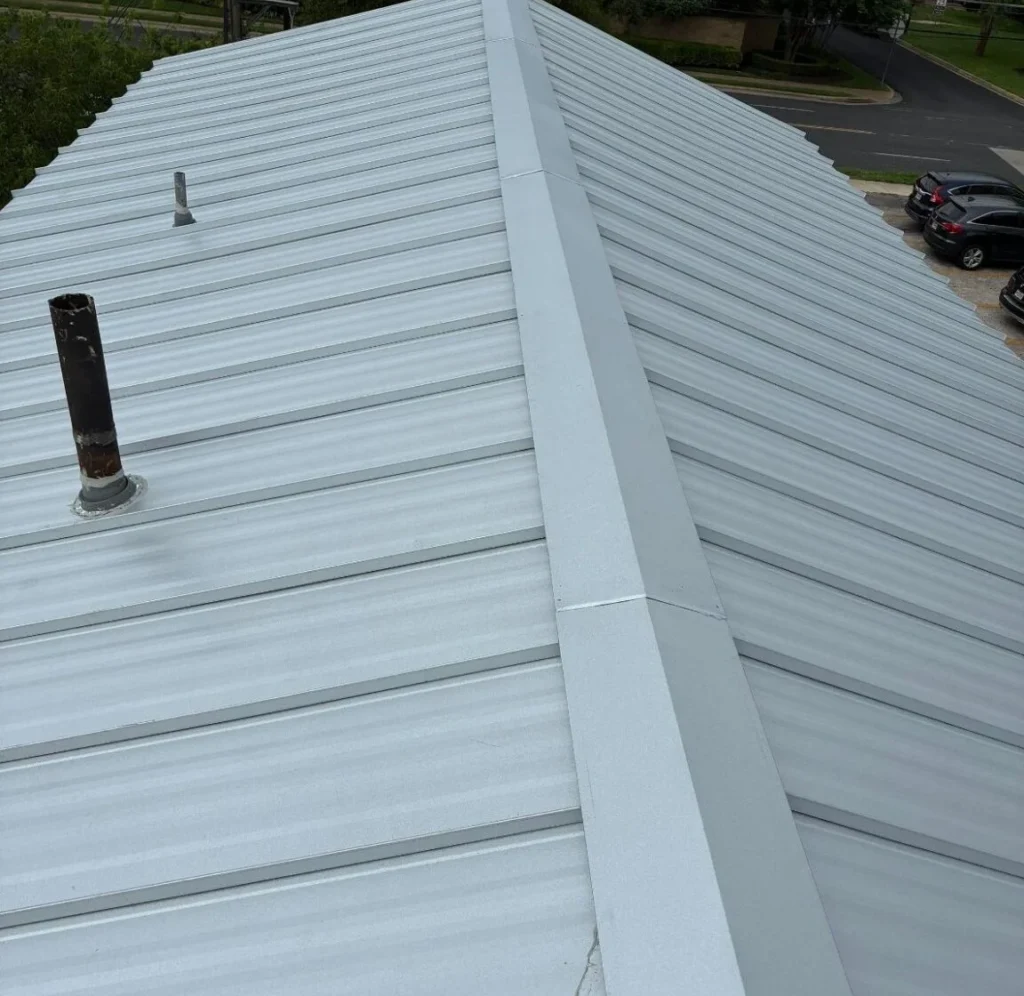 Overhead view of a silver metal roof with a central ridge and multiple standing seams, plus two vent pipes nearby.