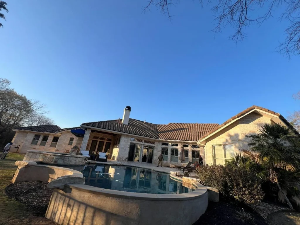 Backyard of a stone-clad house with a curved pool, outdoor seating, and a clear blue sky above.