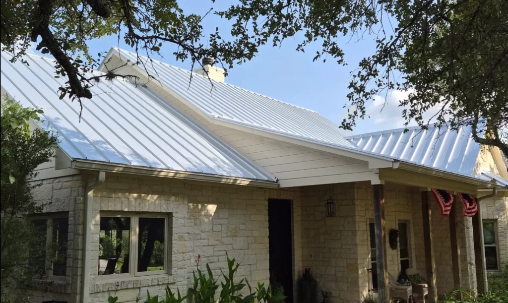 Front view of a stone house with a white metal standing-seam roof and a covered porch; American flags hang at the entrance under a tree.