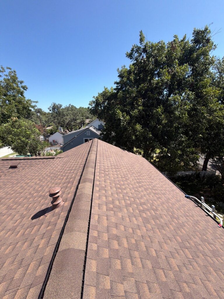Aerial view of brown shingled roof with vents, neighboring houses, and tall green trees against a clear blue sky