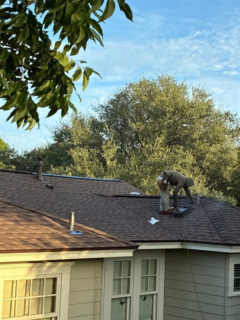 Two workers on a sloped brown shingle roof repairing or installing shingles, with trees and a blue sky in the background.