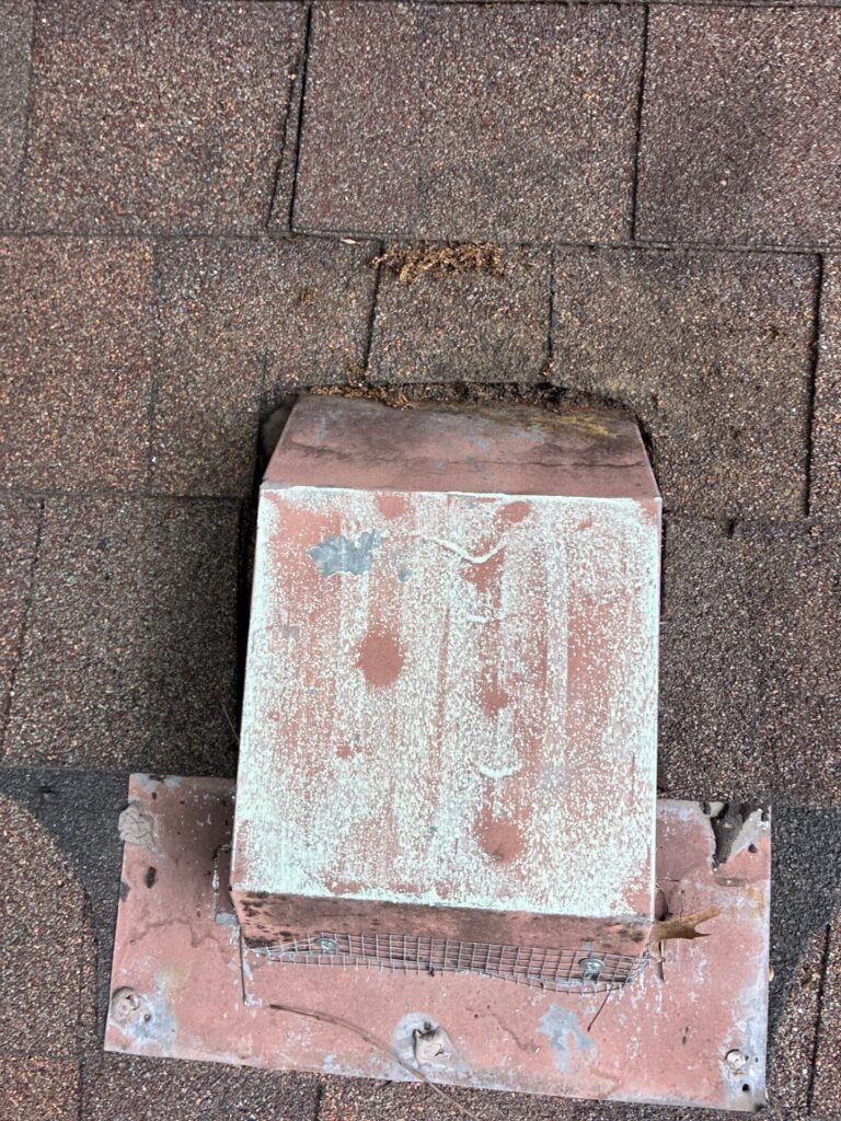 Rusty metal roof vent flashing mounted on brown asphalt shingles, with surrounding weathered shingles and debris.