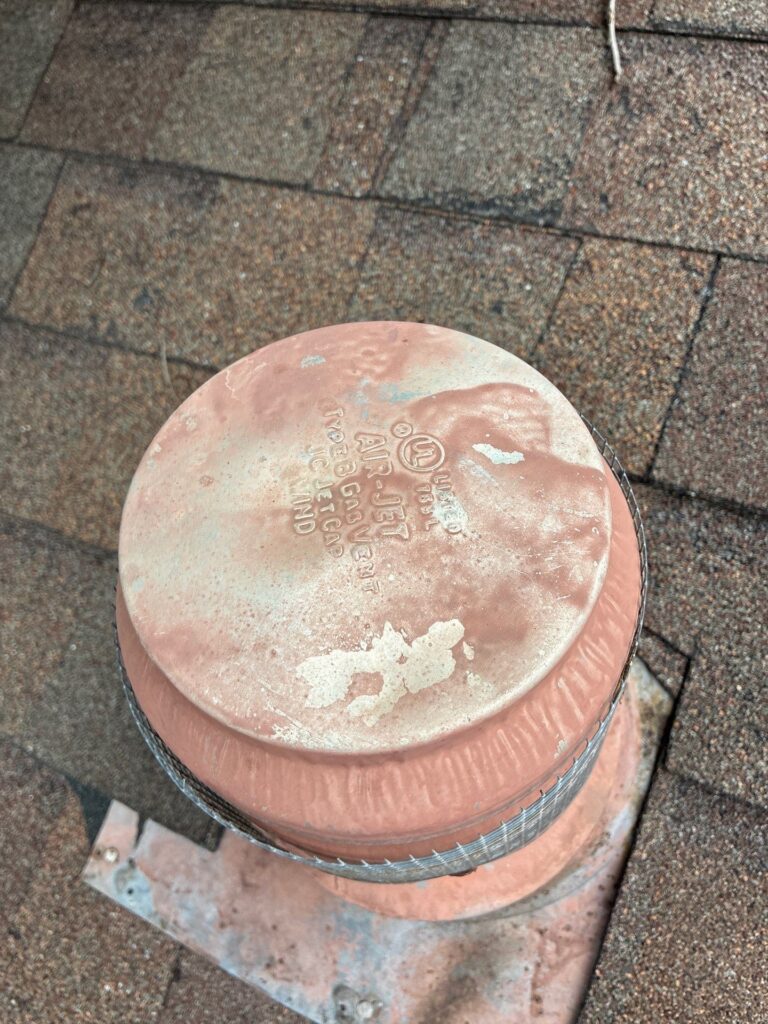 Circular copper roof vent cap on brown asphalt shingles, showing a weathered finish and a mesh-wrapped base edge.