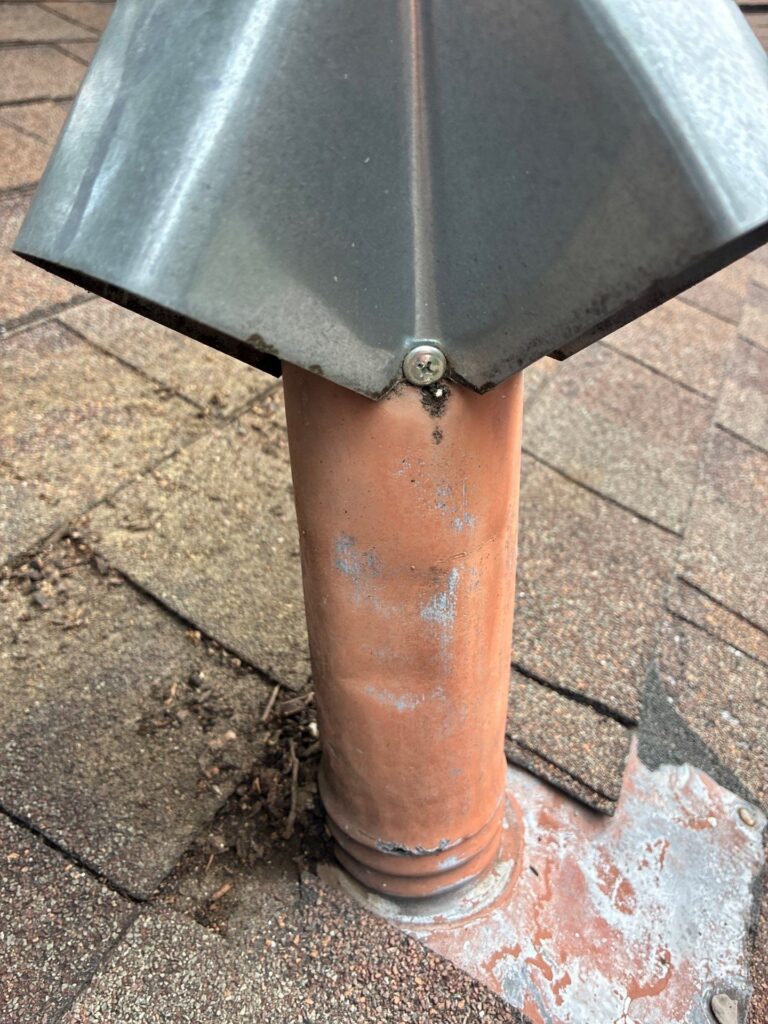 Close-up of a copper vent pipe with a metal flashing cap secured by a screw on a shingled roof surface.