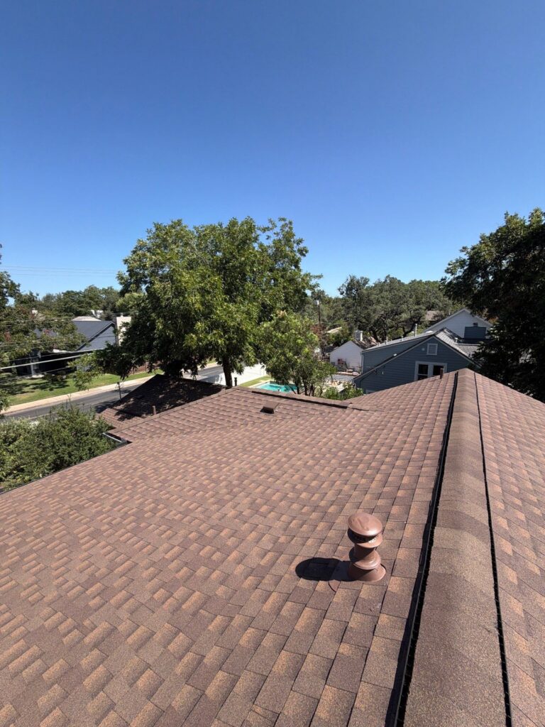 Overhead view of brown shingle roofs, a chimney, and green trees against a clear blue sky.