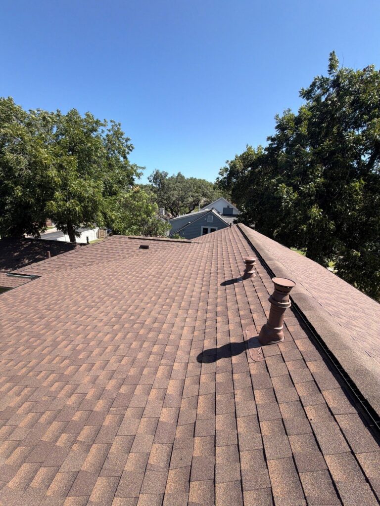Aerial view of a brown shingle roof with two chimney vents, with trees and a clear blue sky beyond.
