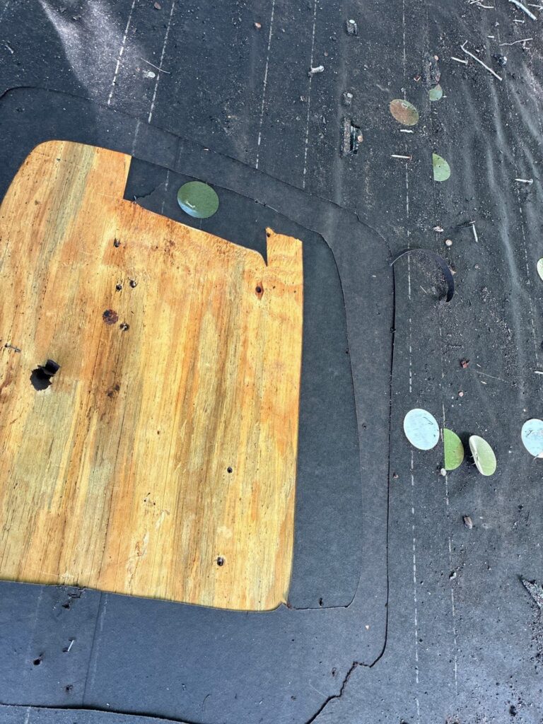 Close-up of plywood edge meeting a black weatherproof membrane with scattered round green discs and small nails, showing wear and cracks on the surface.