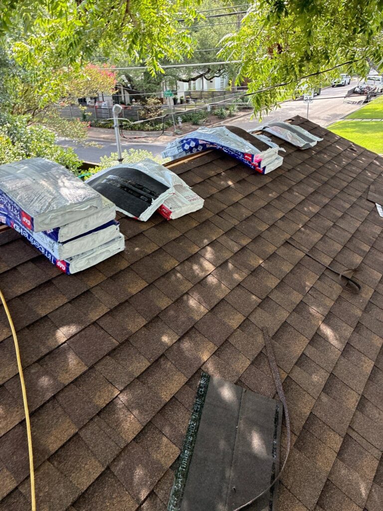 Bundles of roof insulation wrapped in plastic sit on a brown shingle roof, awaiting installation or disposal under a sunny tree-lined street nearby
