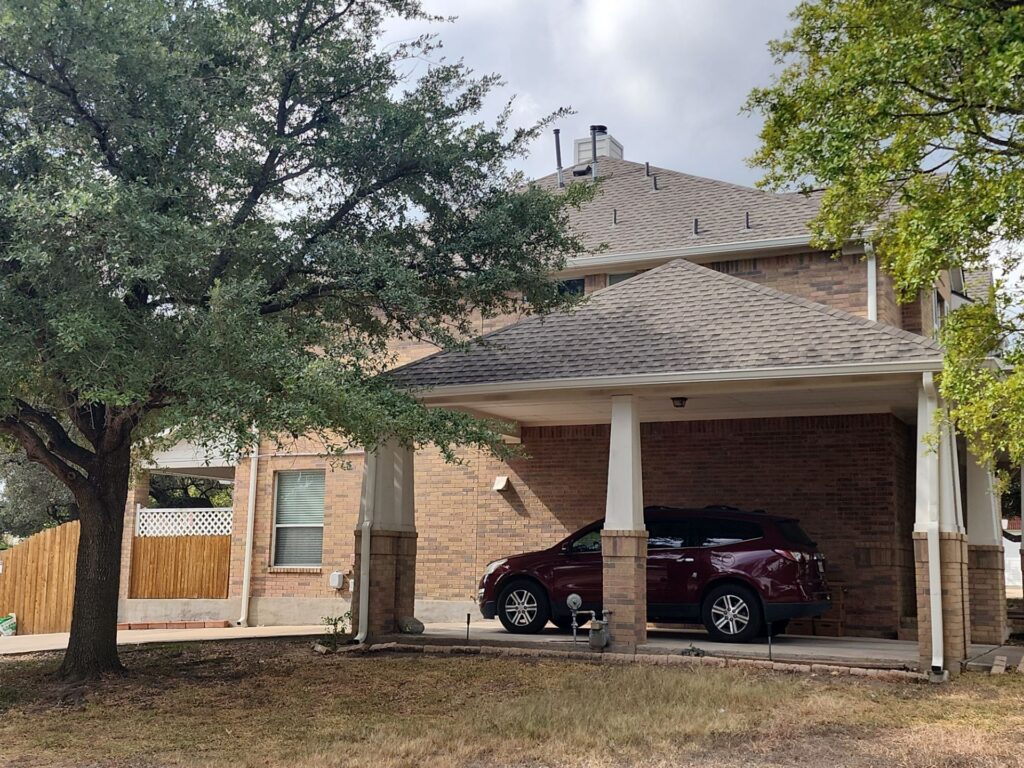 Two-story brick house with a carport and a red SUV parked inside, shaded by a large tree on a cloudy day.