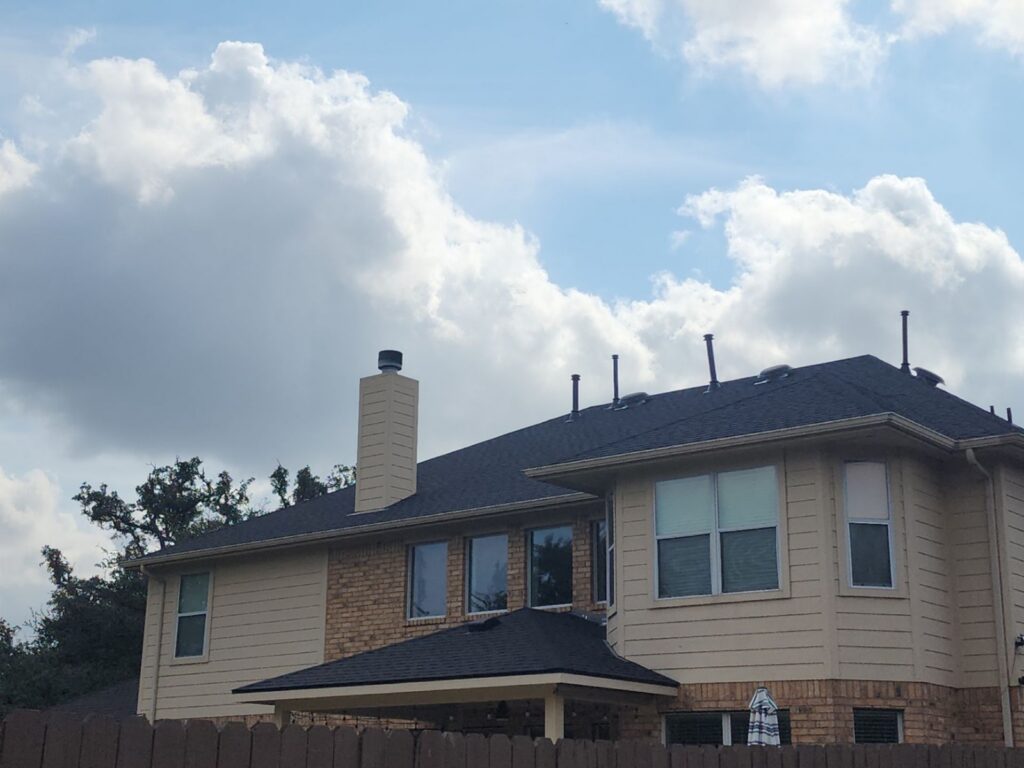 Two-story beige house with brick lower facade, dark roof, chimney, and roof vents under a cloudy blue sky.