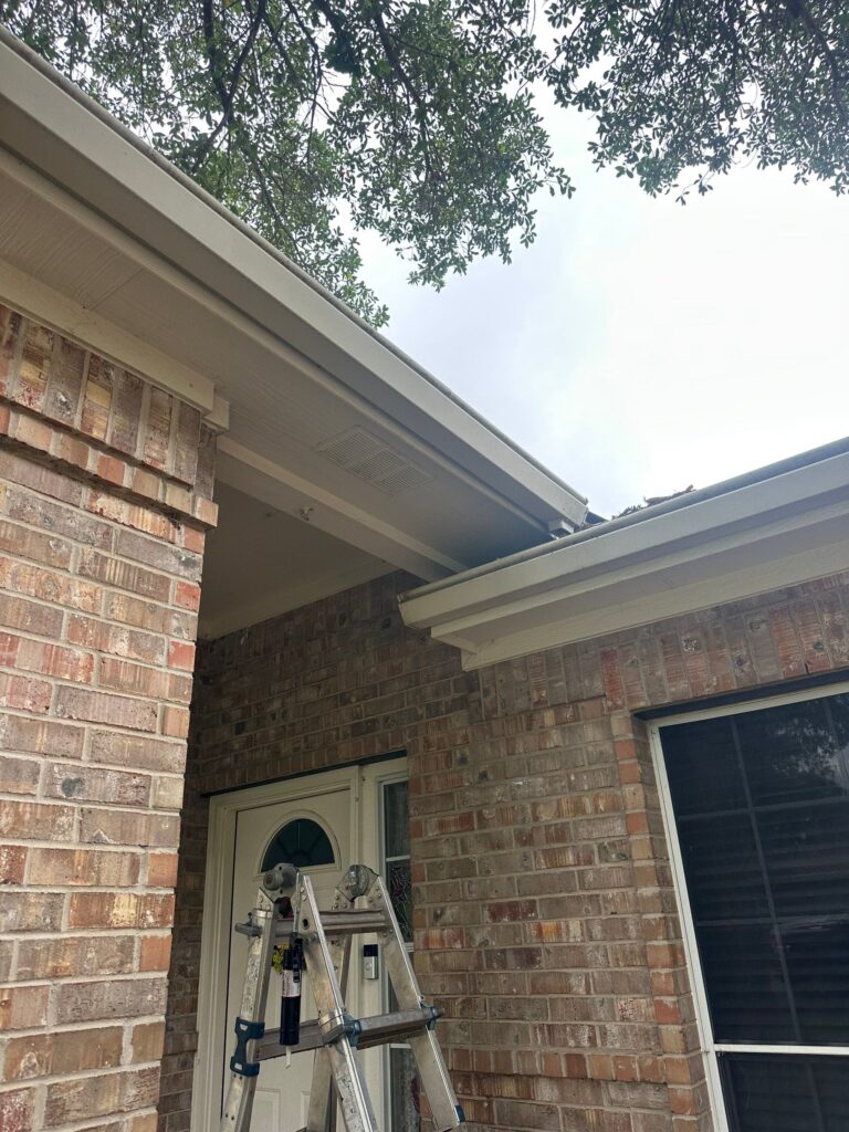White gutters running along the roofline of a brick Austin home, with the soffit, soffit vent and fascia visible and a ladder set up at the entry, on a Driftwood Builders Roofing project