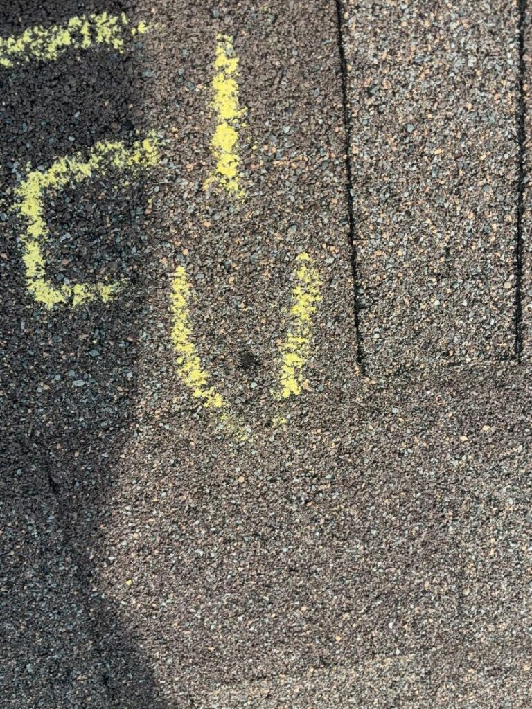 Hail impact marked in yellow chalk on an asphalt shingle, with a darkened strike point visible