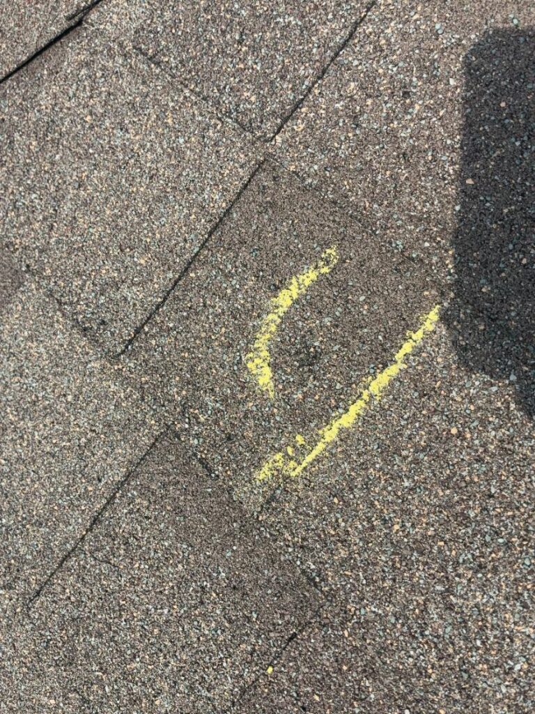 Yellow chalk marks beside a hail impact on an asphalt shingle roof