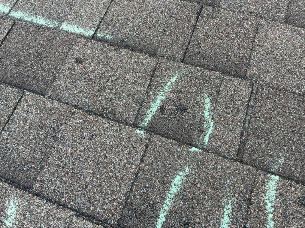 Chalk marks beside darkened hail impact points on weathered asphalt shingles
