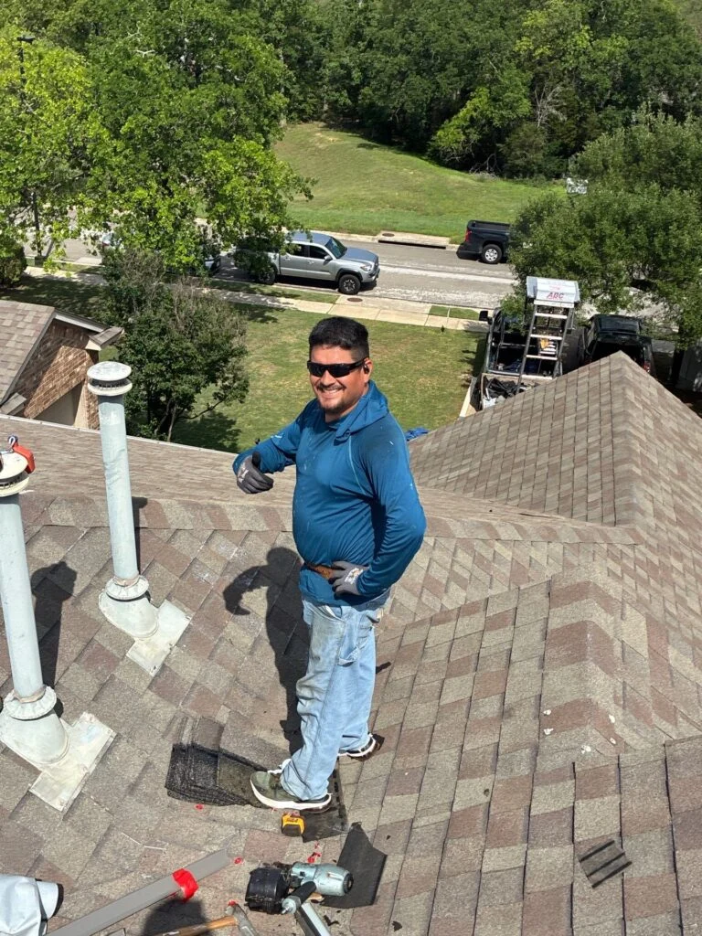 Roofing worker on a sloped asphalt shingle roof wearing a blue long-sleeve shirt and sunglasses, with tools scattered nearby.