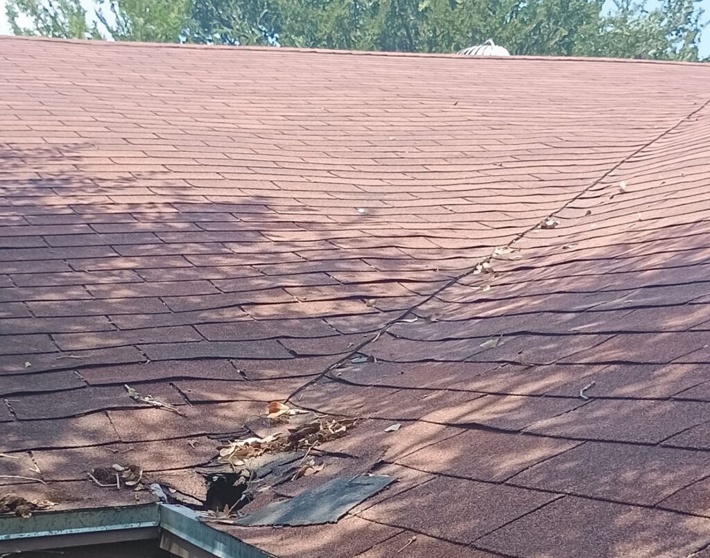 Asphalt shingle roof valley ending at the eave, with torn shingles, packed leaf debris and a hole through to the roof deck at the bottom of the valley